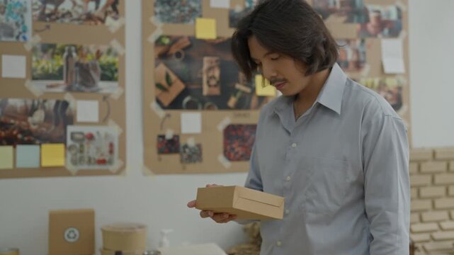Asian adult male portrait in office workspace holding cardboard box, packaging and packaging materials, thoughtful business and creative process with mood board wall in studio workspace