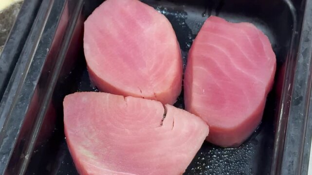 Pink tuna steaks on black tray, supermarket shelf closeup, chilled display of raw fresh fillets arranged in neat rows, subtle.