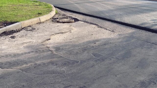 Damaged asphalt curb corner with aggregate, scattered loose asphalt and gravel along cracked pavement next to grassy verge.