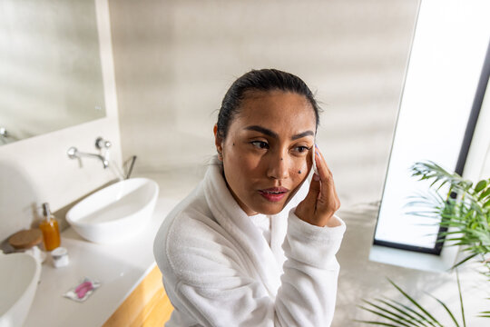 Asian female wearing white bathrobe standing at bathroom sink removing makeup with cotton pad