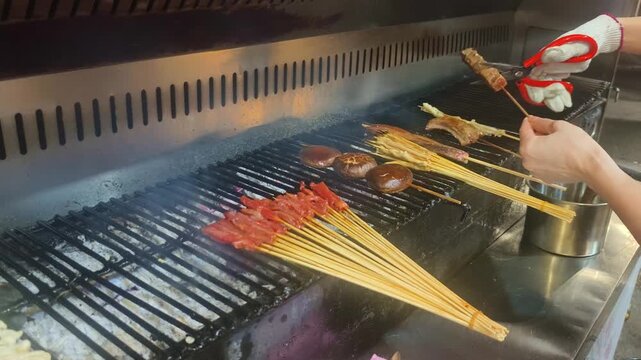 A chef grills skewers of marinated meat, mushrooms, and other ingredients over charcoal on a commercial barbecue grill, with smoke rising from the fire