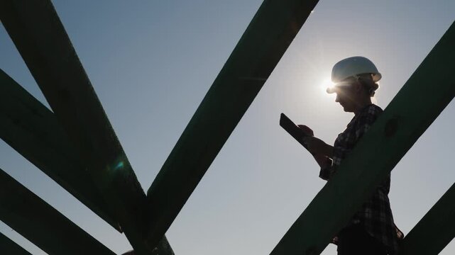 Woman builder inspects construction project using a digital tablet at sunset, checking house framing against blue sky with bright sun.