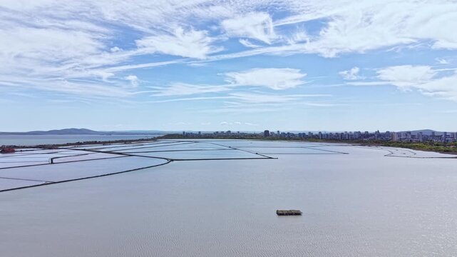 Water covers the area with salt pans arranged in lines. In the distance, a city skyline is visible under a clear blue sky. A boat floats in the water.