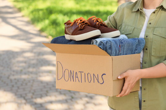 Woman holding donations box with folded clothes and shoes outdoors, charity clothing drive, community support, giving away used apparel for recycling and helping people in need
