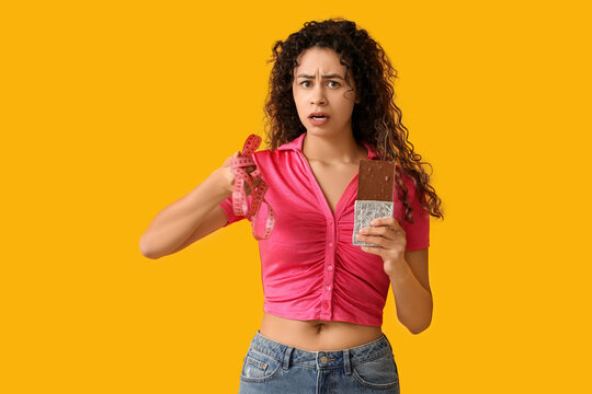 Beautiful young African-American woman with sweet chocolate bar and measuring tape on yellow background