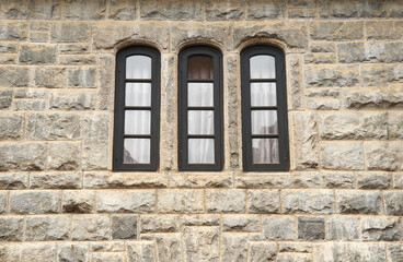 Fototapeta premium three narrow arched black windows on rustic stone wall, side view of vintage residential building with copy space