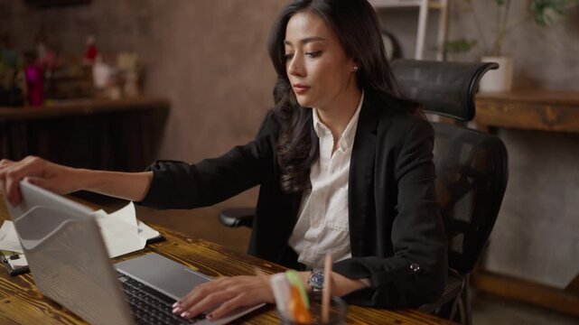 Female office worker seated at desk, focused on laptop and paperwork in home business office, professional woman working on computer in modern workplace interior
