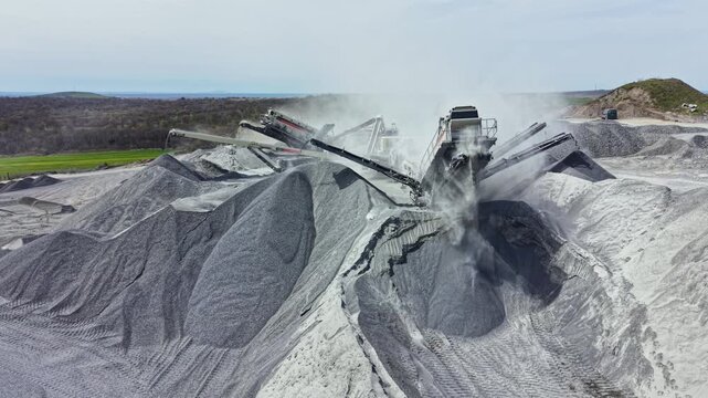 A large machine works in a granite quarry, crushing and processing rock while dust rises in the air during the day.