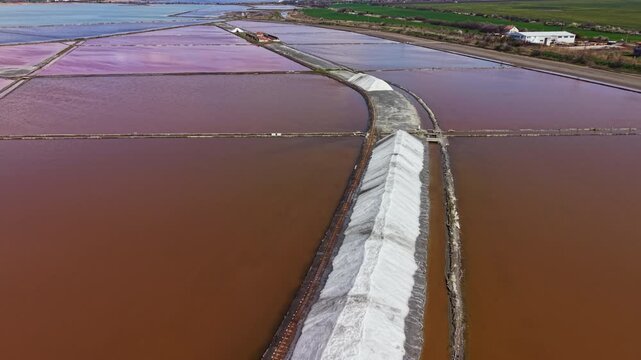 Salt is harvested in shallow ponds arranged in a grid. Workers manage water levels and collect salt at a salt production site near the sea. The landscape shows various water colors.