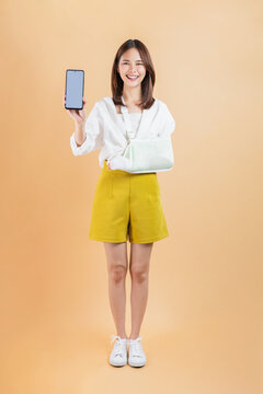 Asian woman broken arm wrapped holding smartphone mockup of blank screen isolated on beige color background