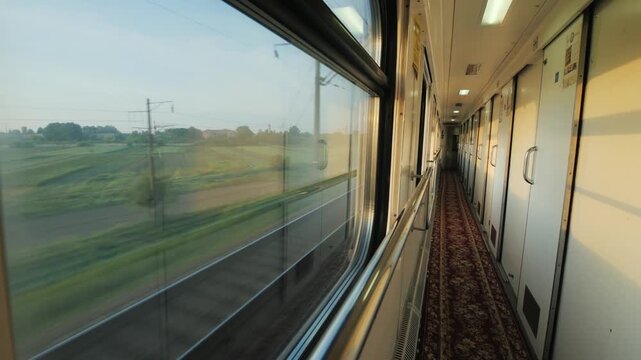View from moving train window looking at green fields and empty railroad track at sunrise, long corridor with closed doors and carpet inside train wagon, cinematic motion footage.