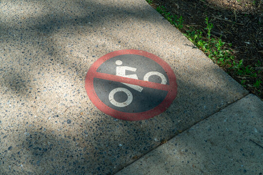 No Bicycles No Skateboards Sign on Textured Concrete Pavement Next to Dirt with Green Weeds, Emphasizing Prohibited Activities in Public Outdoor Space with Sunlight and Shadow