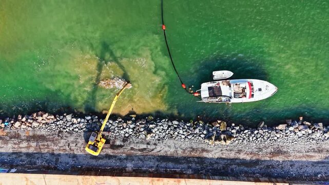 A crane is working on the shore while a boat is anchored nearby. Workers are engaged in maintenance tasks in the water along the coast in broad daylight.