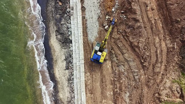 Workers use heavy machinery for construction along the shoreline where land meets water. The site has dirt, rocks, and concrete blocks for building.