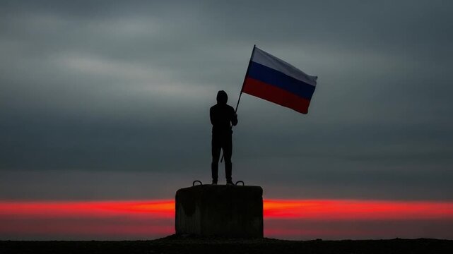 Silhouette of a young man holding a Russian flag standing on a concrete block against a dramatic sunset sky