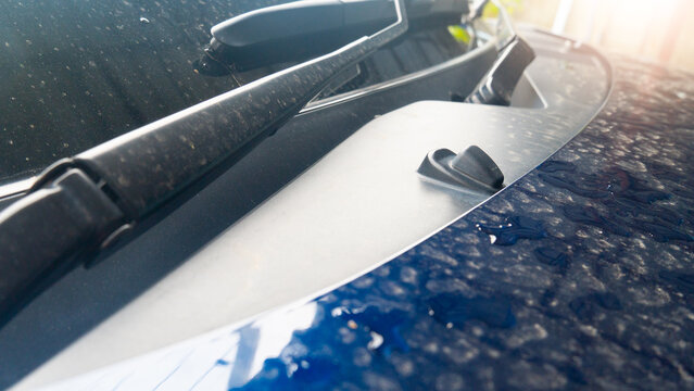 Close-up of a blue car hood covered in water droplets and dust. The image shows the windshield wiper arm and a black washer nozzle. Sunlight reflects on the wet, textured surface of the vehicle.