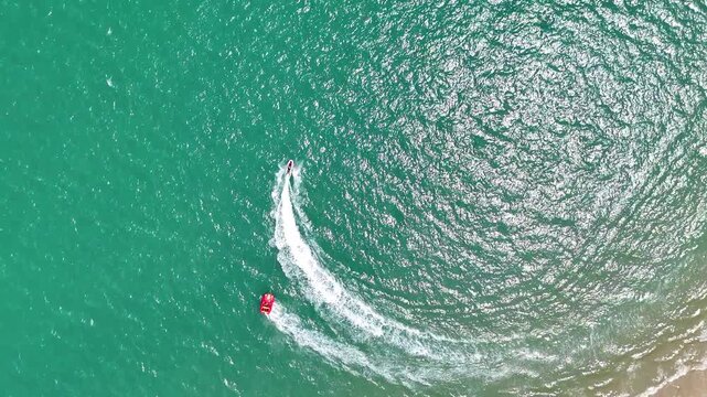 Aerial view drone shot of a jet ski pulling a red banana boat with tourists, creating white water waves near the sandy beach