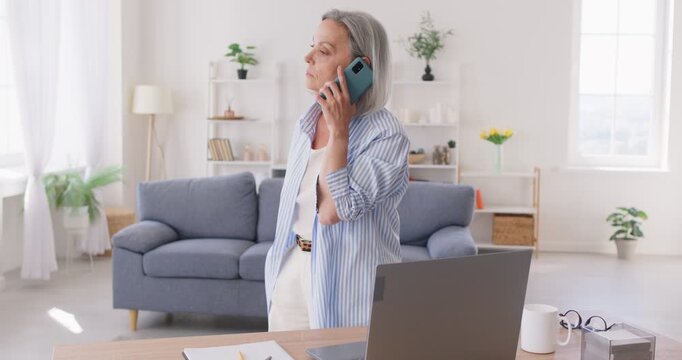 Senior businesswoman answers phone call while working at home. Busy old gray haired business woman standing by desk, typing on laptop picks up cellphone and starts walking back and forth while talking