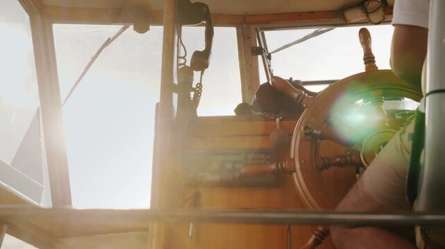 Man steering boat at sunset from inside cabin, hands on wooden wheel.