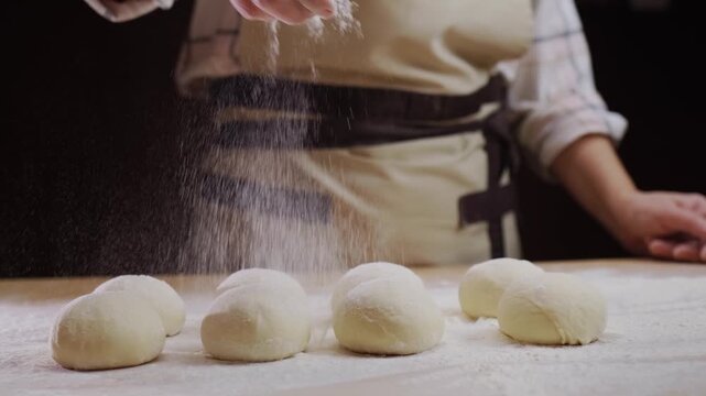 Baker dusting raw dough balls with flour on a wooden table preparing for baking.