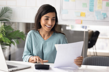 Fototapeta premium Latina accountant woman sits at office desk holding paper documents, smiling while using calculator, reviewing financial or business data with satisfaction, performing accounting or bookkeeping tasks