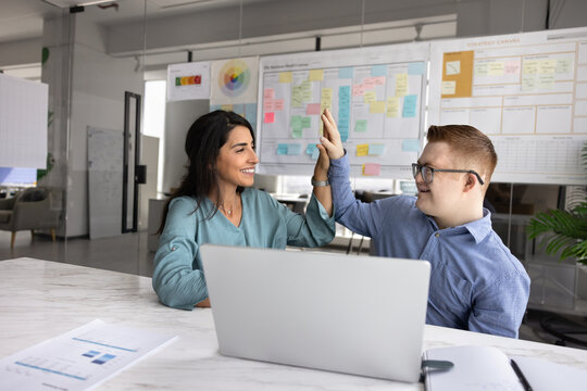 Two coworkers, man with Down syndrome and woman, exchanging high five gesture, celebrating achieved goals, accomplished joint task, seated at desk with papers and laptop. Teamwork and project success