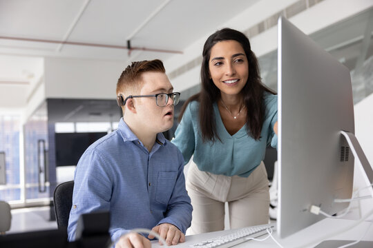 Brazilian woman colleague helps with online task to male employee with Down syndrome, reviewing project data, studying new corporate software or platform together, working in modern coworking office