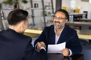 Fototapeta na wymiar Smiling middle-age hr manager holds resume paper handshakes with male applicant across office table. Business agreement, partnership approval, hiring, commercial deal closure, successful negotiations
