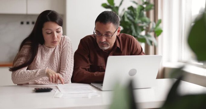 Couple sitting at kitchen table handling household finances, wife calculating expenses while husband paying bills using online banking on laptop. Family budgeting, financial planning and management