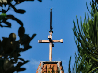 Obraz premium Copper christian cross atop a church roof framed by green tree branches against a clear blue sky