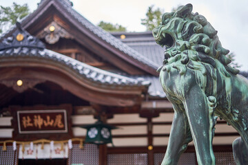 A weathered bronze komainu (guardian lion-dog statue) with green patina stands prominently in the foreground right, mouth open in a protective stance. Behind it, a traditional Japanese shrine © Andrius