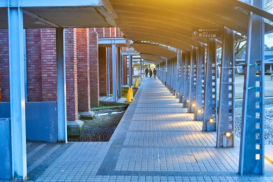 A covered pedestrian walkway featuring modern industrial architecture with red brick columns on the left side and metal support pillars on the right. The pathway is paved with rectangular tiles and