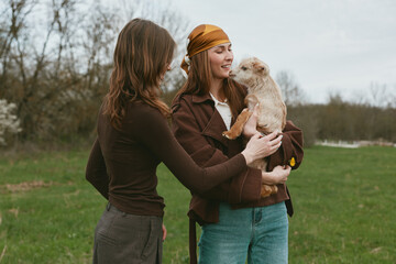 Fototapeta premium Outdoor lifestyle scene with two women smiling as one holds a goat kid, softly lit by natural light, casual jackets, grassy meadow background, carefree connection, warm tones, candid moment.