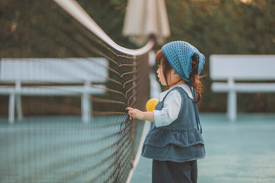 Cute Asian toddler girl holding an orange ball on a pickleball court at sunset. Happy child in denim outfit learning sports, promoting active lifestyle, wellness, and childhood development