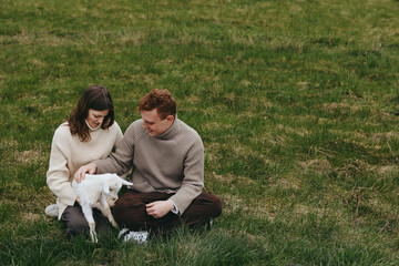 Fototapeta premium Lifestyle couple gently pets a white goat kid in an outdoor grass field, cozy sweaters, natural candid moment, warm bonding and calm affection, a simple rural scene of care and joy.