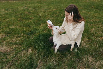 Fototapeta premium A woman sits on grassy ground feeding a young goat kid with a bottle in an outdoor field, capturing a tender lifestyle moment of care, bonding, and simple countryside joy.