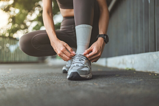 Close up of an Asian fitness woman tying her sneaker shoelaces while crouching down on an outdoor park path before morning exercise. Healthy active lifestyle, running preparation, wellness