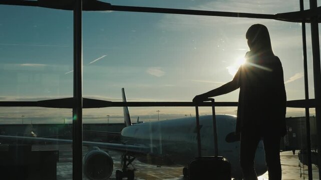 Silhouette of a woman with luggage watching airplane through airport window at sunset.