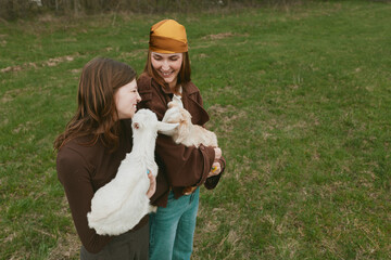 Fototapeta premium Lifestyle outdoor scene with two women in a grassy field, goat kid held between them, laughter and warmth, casual clothing, natural colors, affection and friendship shown, calm rural moment, joyful