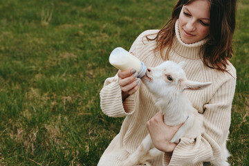 Fototapeta premium Cheerful woman outdoors, feeding a white goat kid with a bottle, cozy sweater, sunlit field, candid moment, gentle interaction, natural tones, pastoral scene, relaxed mood
