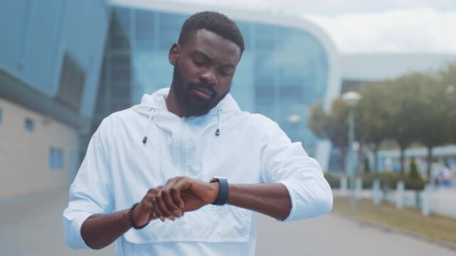 Male pedestrian in relaxed attire inspects his smartwatch amidst cityscape environment during late afternoon