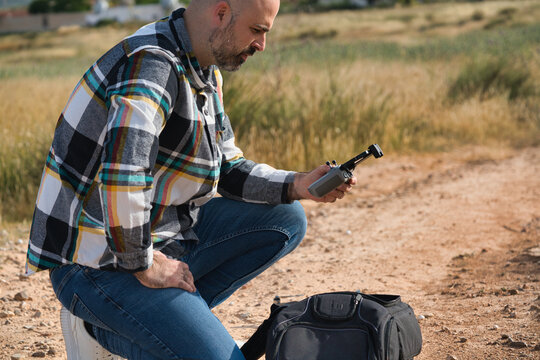 A man on a dirt road, examining a drone to practice flying.