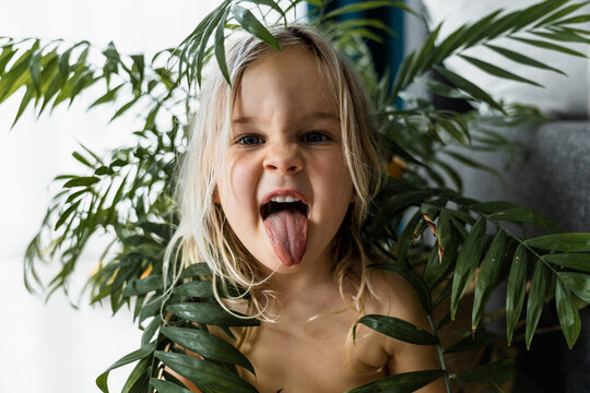 Funny toddler girl sticking tongue out among tropical houseplants