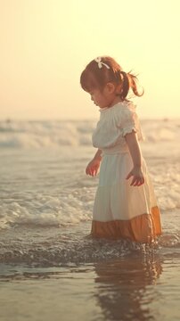Adorable Asian toddler girl in a white dress walking playfully in the sea waves at the beach during a warm golden sunset. Happy childhood, family summer vacation, carefree weekend getaway