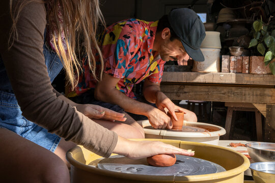 Young man and woman spinning clay on wheel in window light