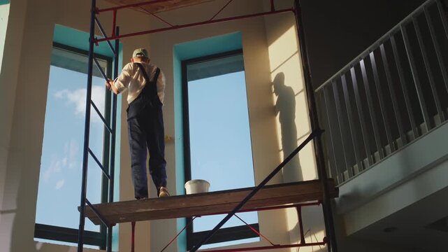 A male worker in work clothes carefully paints the wall white while standing on scaffolding, slow motion footage.