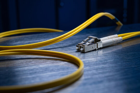 Elegant editorial close-up of a matte canary-yellow optical patch-cord arching over horizontally brushed steel, illuminated by crisp 170-degree backlighting and sharp rim light.
