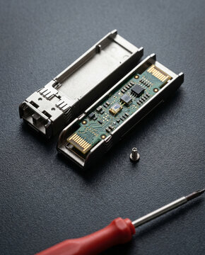 Technical teardown flatlay of a disassembled SFP+ module, revealing an internal miniature green circuit board with micro gold solder points on a dark fine-grained textolite surface.
