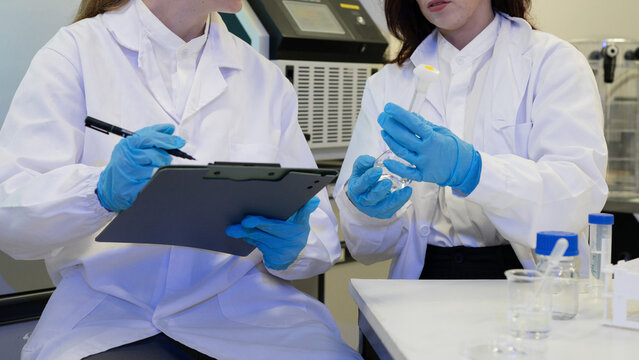 Two Female Scientists in Lab Coats Conducting Experiment with Clipboard and Flask in Laboratory