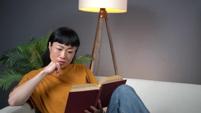 Focused Asian Woman Reading Book at Home on Sofa and Deeply Thinking about Story Plot or Education Content